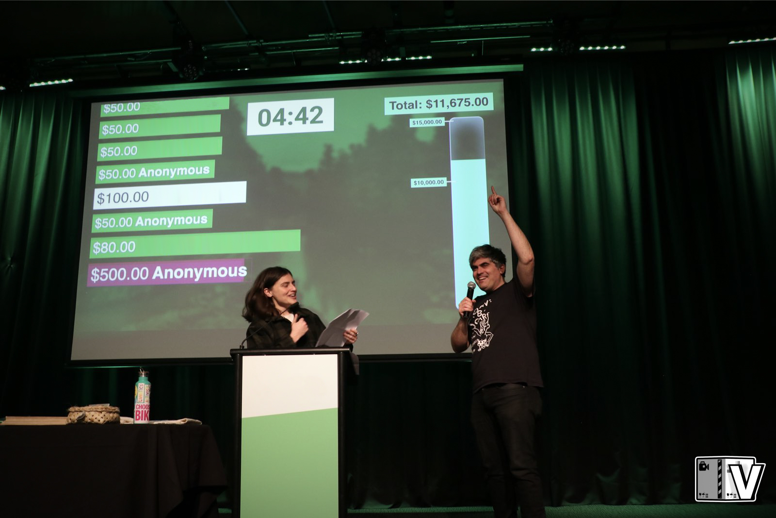 Chlöe Swarbrick MP and Mayor Aaron Hawkins hosting a fundraising event for the Green Party of Aotearoa New Zealand. Aaron points at the screen which has a live ticker of pledges and a thermometer showing progress towards their goal, while Chlöe reads off a paper into a lecturn microphone.
